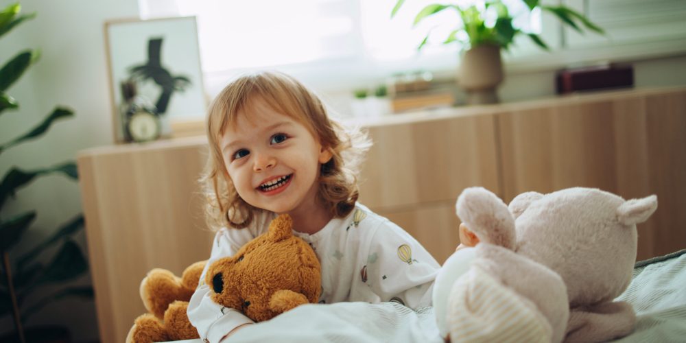 Portrait of adorable baby playing in the bedroom. She is smiling and holding stuffed animal toys. There are plants around the room.